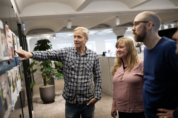 En man i ett kontor pekar på en anslagstavla, medan två kollegor - en annan man och en kvinna - tittar på.