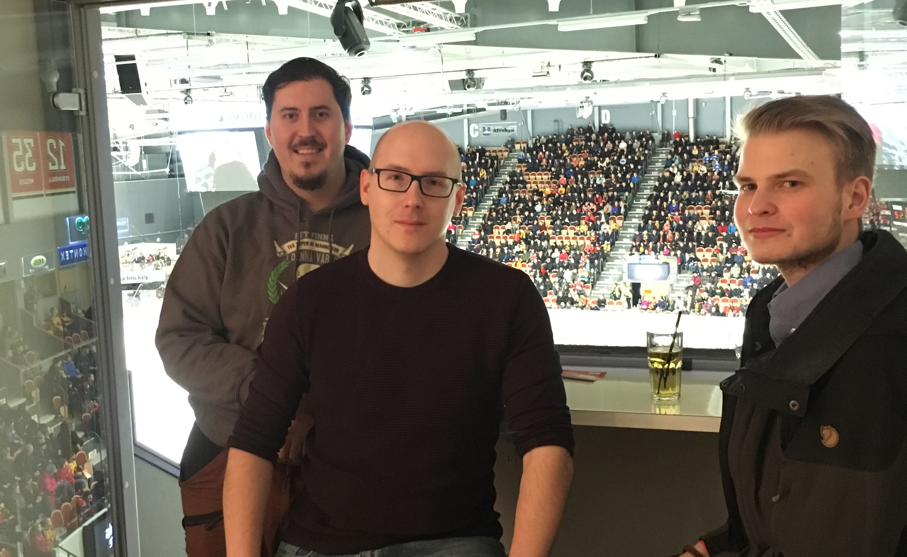 Bilden föreställer tre män som står i en utsiktsloge i en hockeyrink. I bakgrunden syns läktare och publik.
