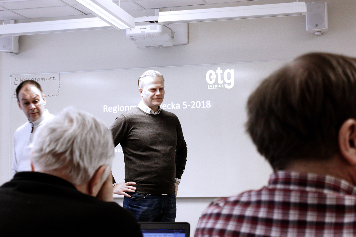 Bilden föreställer en man som står med händerna på höfterna framför en whiteboard där ordet ETG projicerats med hjälp av en projektor. I förgrunden syns ryggarna på två åhörare.
