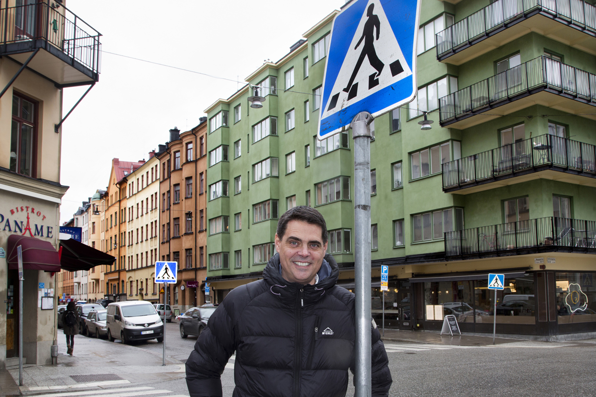 Bilden föreställer en leende man invid en skylt för ett övergångsställe i en vägkorsning.