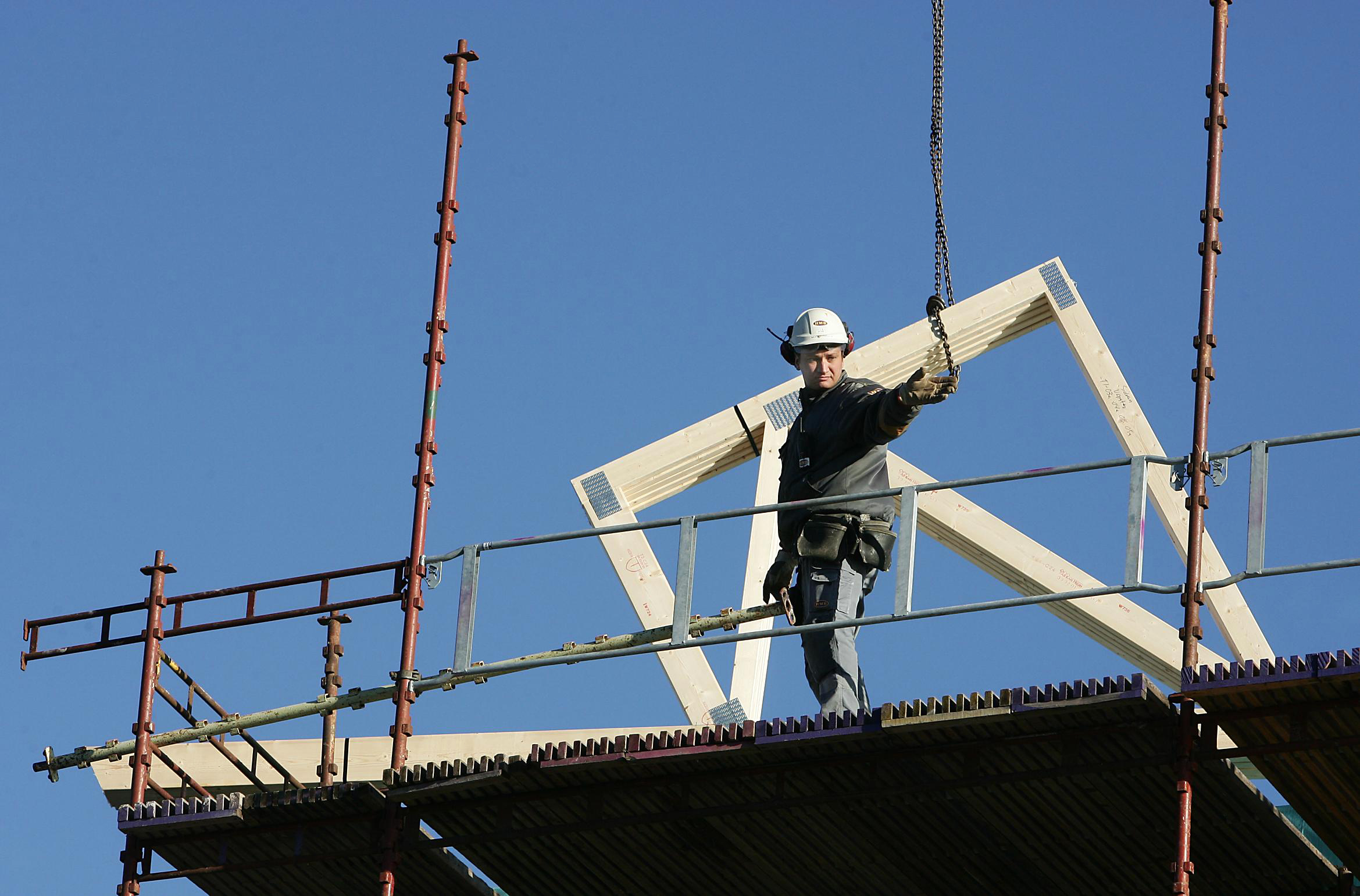 Bilden föreställer en person i vit bygghjälm som står på en byggnadsställni8ng och sträcker ut en hand. Bakom honom syns en träram som lyftas i en kedja.