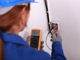 En elektriker i blå kläder och hjälm mäter spänning i ett eluttag.