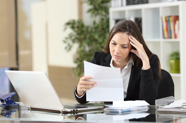En person sitter vid ett skrivbord med en laptop, och läser bekymrat från ett papper.