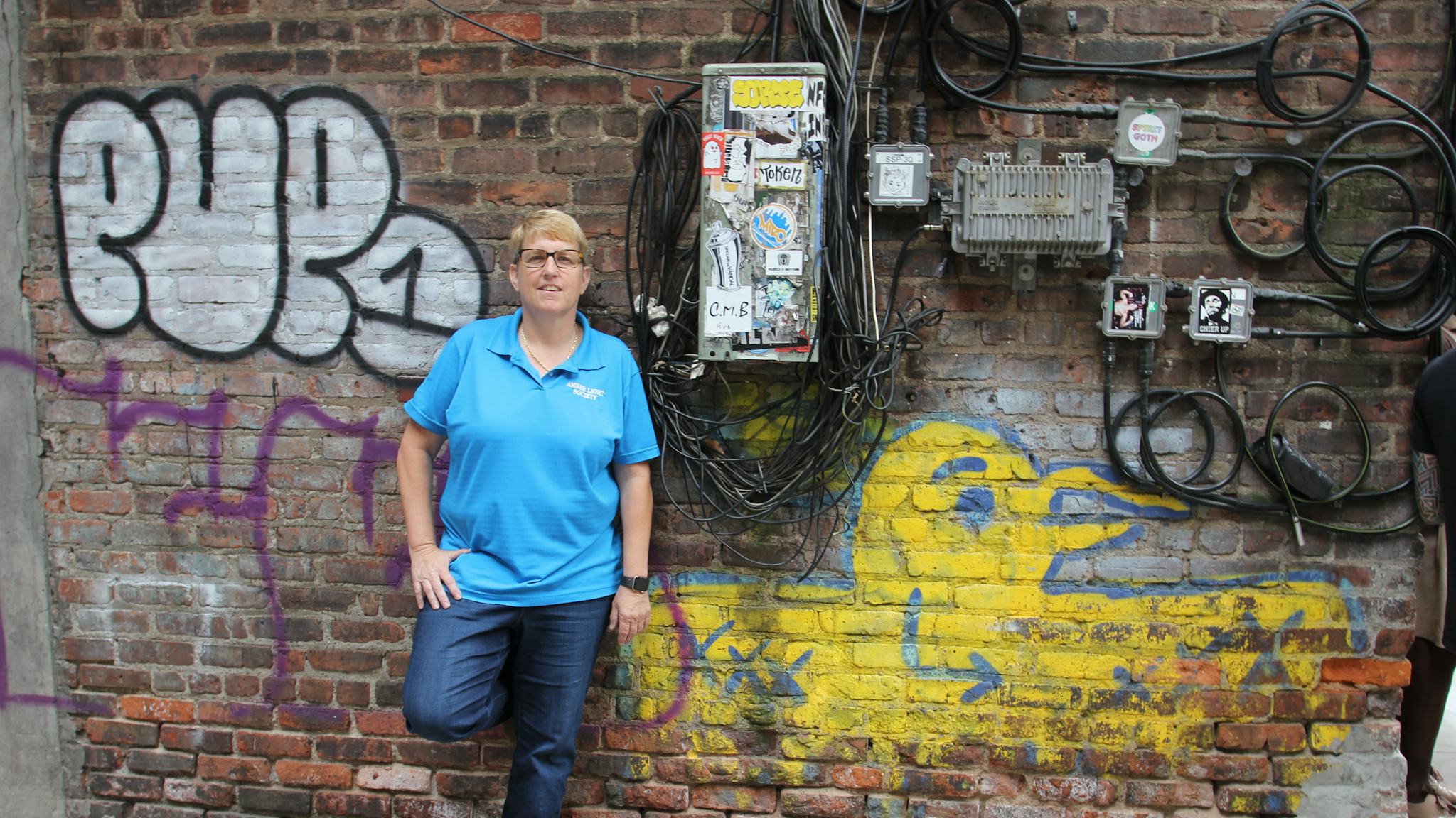 Erin står lutad mot en tegelvägg ful av graffiti, klädd i blå t-shirt.