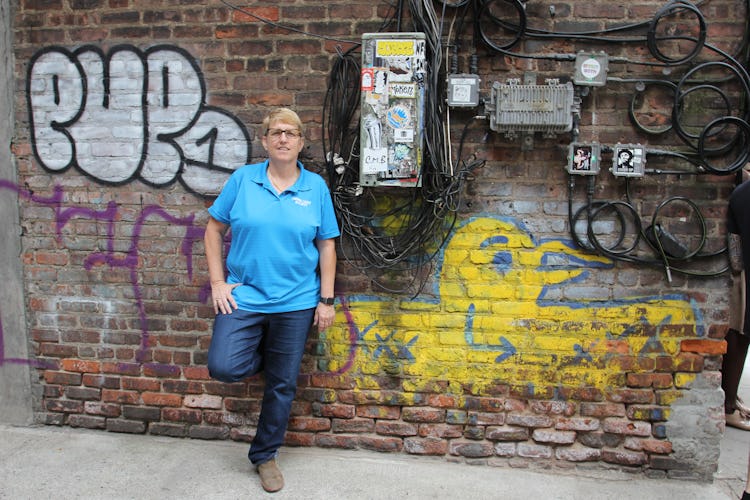 Erin står lutad mot en tegelvägg ful av graffiti, klädd i blå t-shirt.