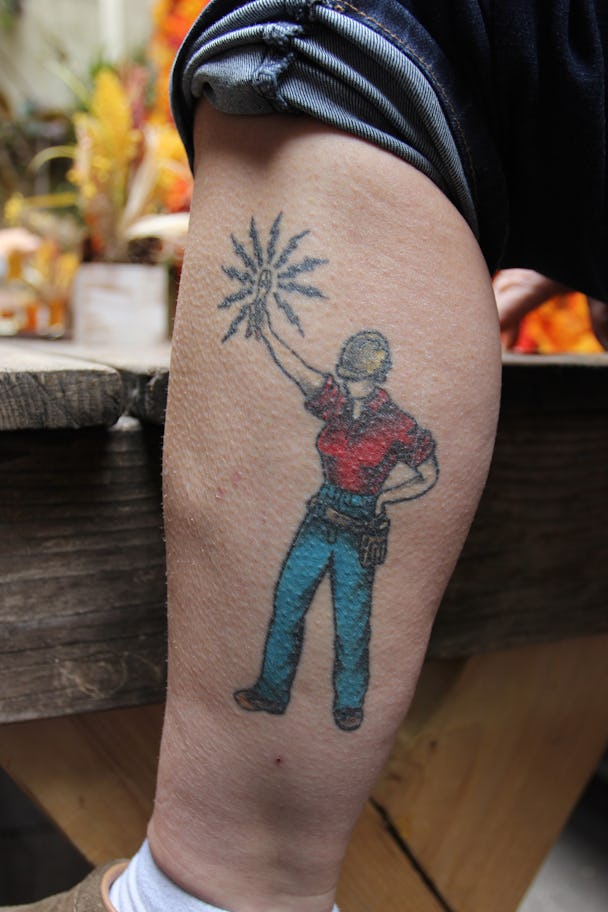 Tatueringen på Erins vad. En kvinnlig elektriker i röd t-shirt och gul skyddshjälem håller upp en el-sprakande tång.