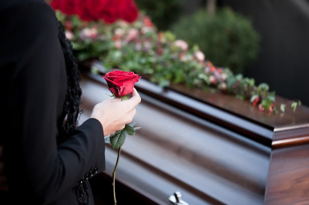 En träkista med blommor på. I förgrunden en person i svarta kläder som håller i en röd ros.