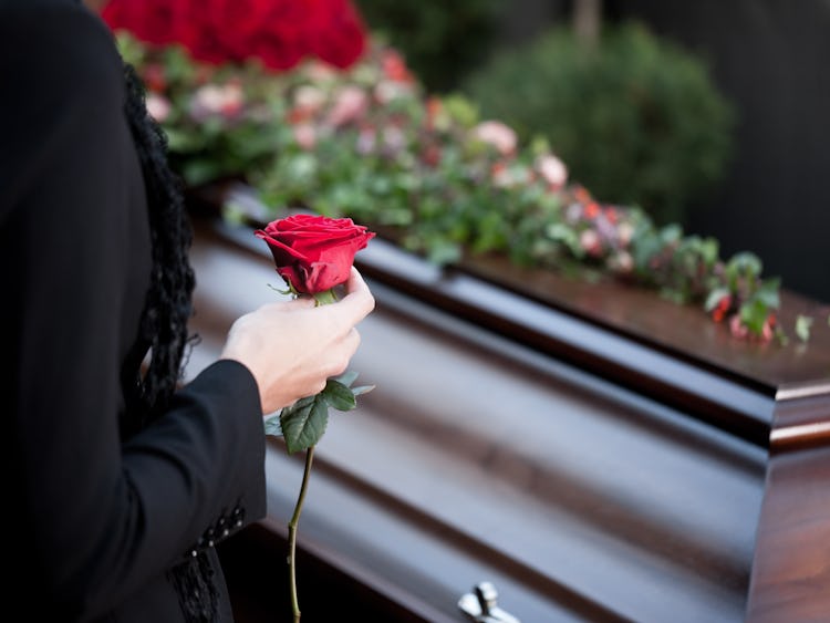 En träkista med blommor på. I förgrunden en person i svarta kläder som håller i en röd ros.