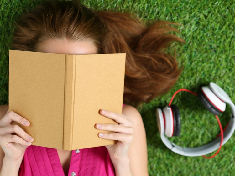 En person logger I gräset och läser en bok, med ett par hörlurar bredvid sig