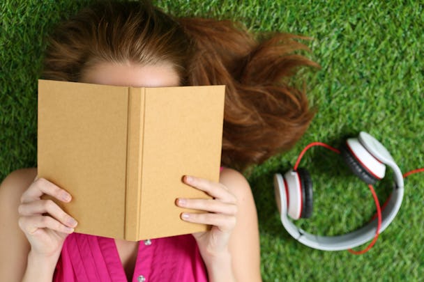En person logger I gräset och läser en bok, med ett par hörlurar bredvid sig