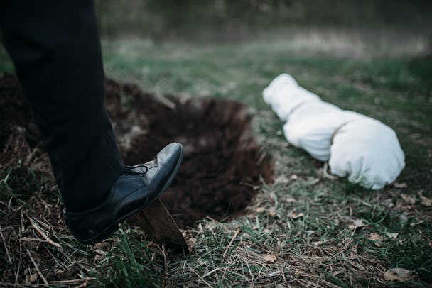 Foten på en person som gräver en grav med en spade. Bredvid gropen ligger ett långt vitt bylte