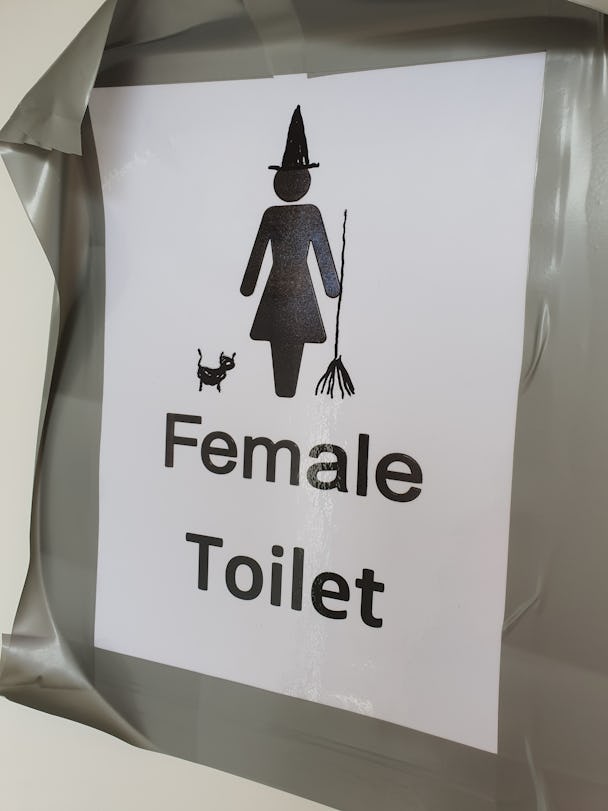 En toalettskylt där den stiliserade kvinnosymbolen har ritats om till en häxa md hatt, kvast och katt.