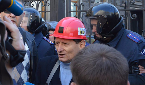 Mykhailo Volynets i röd bygghjälm omgiven av reportrar och personer i kravallutrustning.
