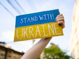 En hand håller upp en skylt med Ukrainas flagga, samt texten: "Stand with Ukraine."