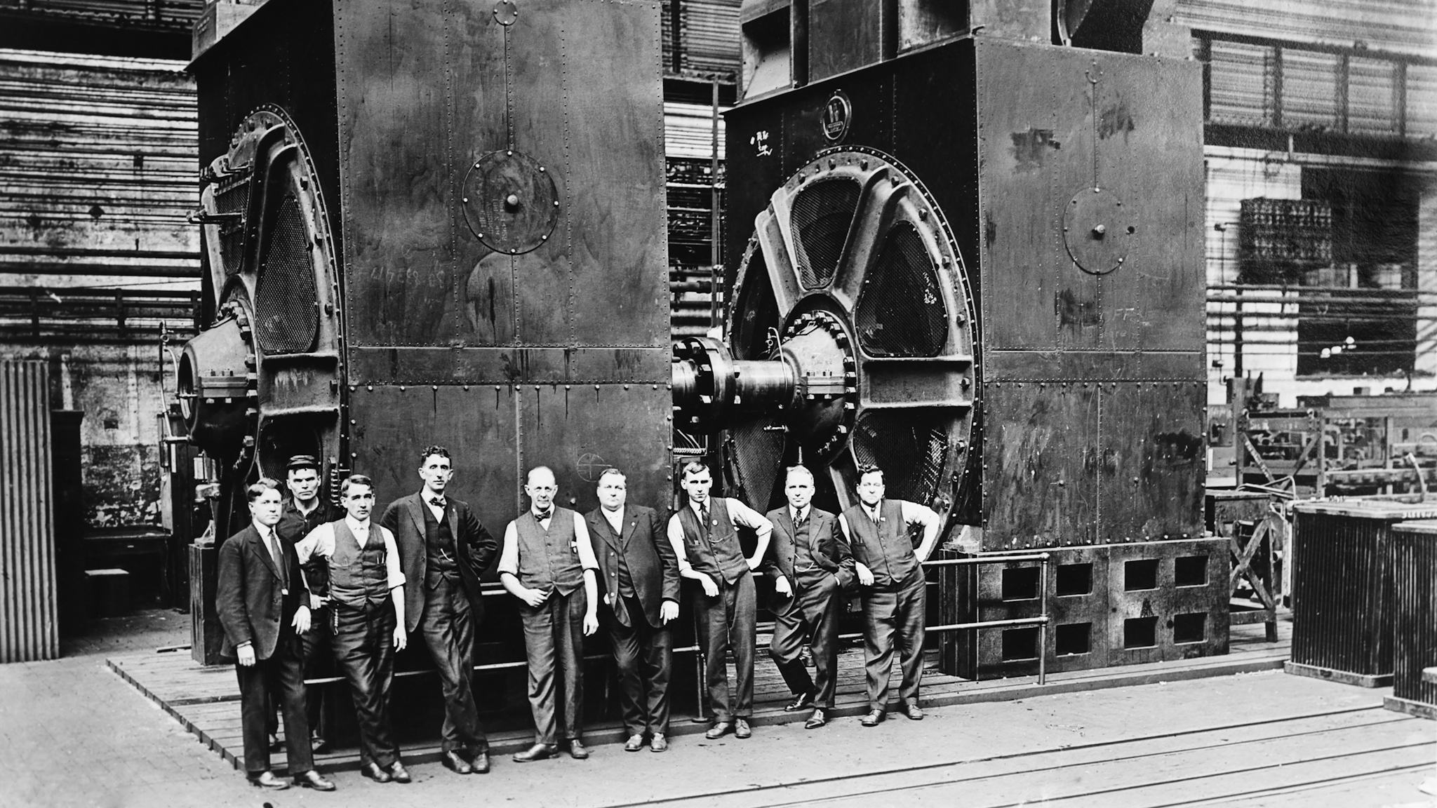 Nio kostymklädda män poserar framför två massiva elektriska generatorer i en fabrik. Svartvitt foto, cirka 1920.