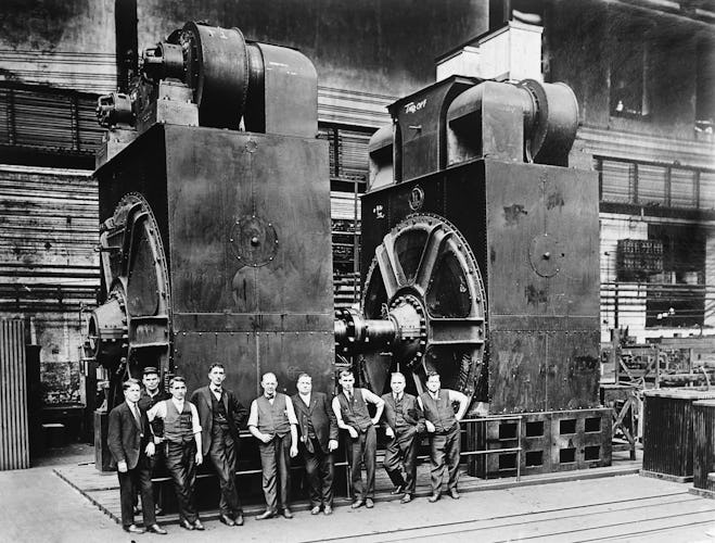 Nio kostymklädda män poserar framför två massiva elektriska generatorer i en fabrik. Svartvitt foto, cirka 1920.