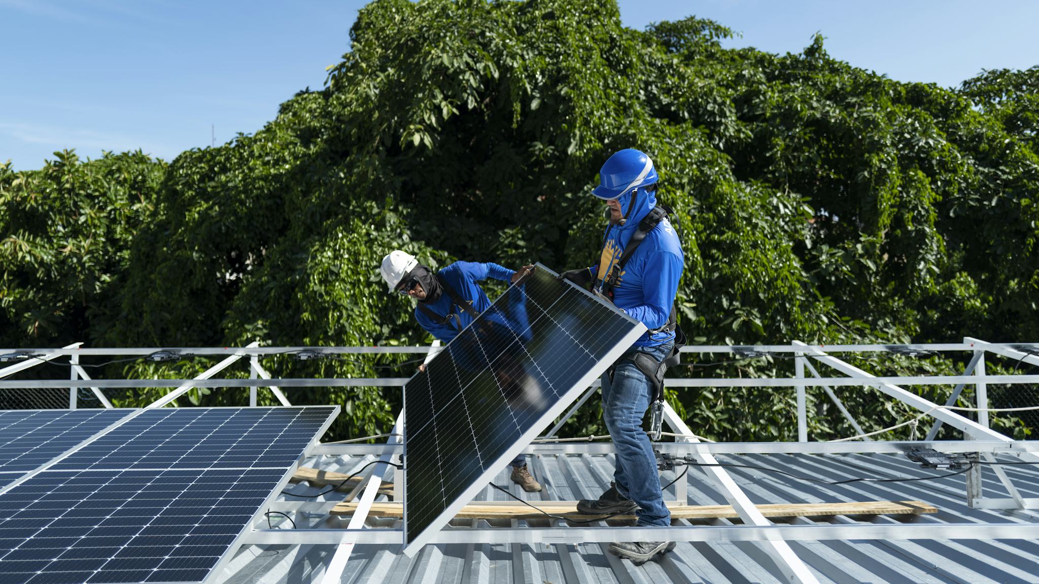 Två arbetare som installerar solpaneler på ett metalltak med träd i bakgrunden.