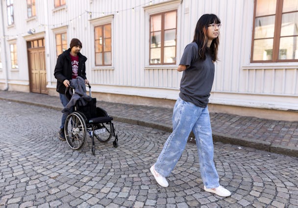En kvinna går på en kullerstensgata medan en man i rullstol följer efter.