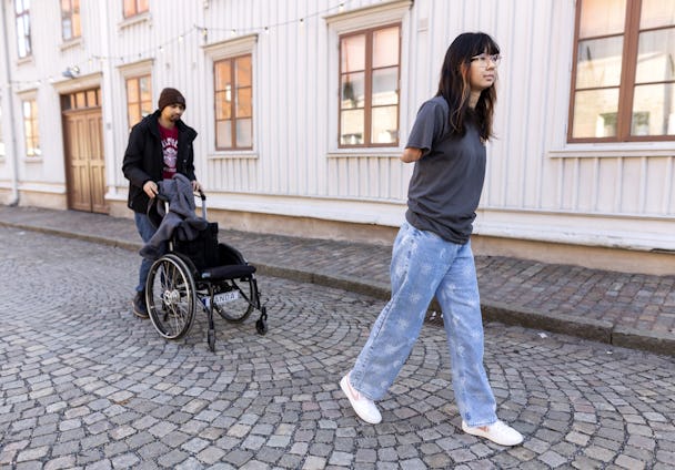 En kvinna går på en kullerstensgata medan en man i rullstol följer efter.