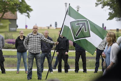 En man håller en grön och vit flagga med en svart symbol under ett utomhusmöte.