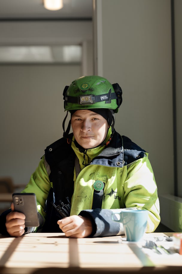 En person med en grön skyddshjälm och gul jacka sitter vid ett bord och håller en smartphone.