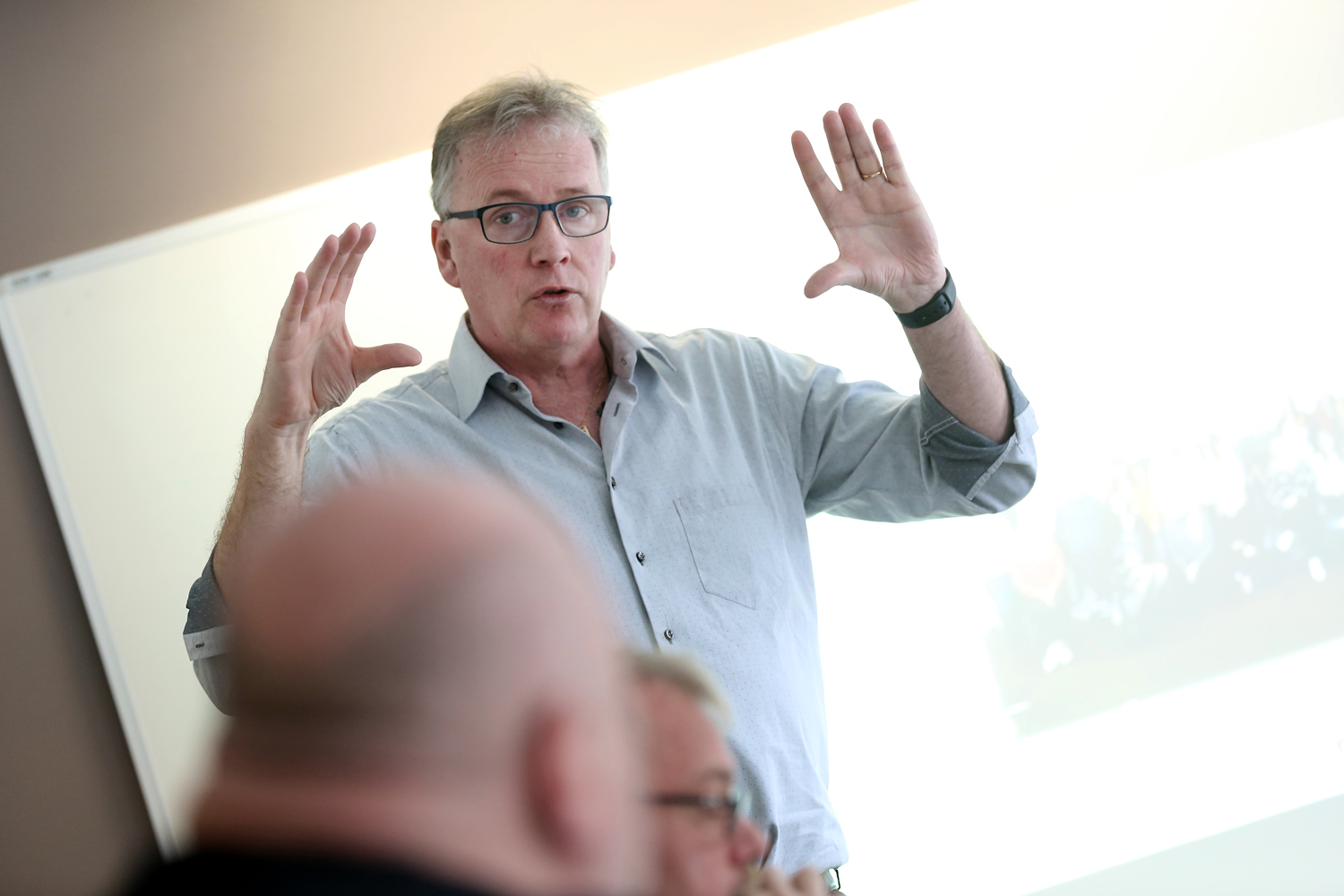 Bilden föreställer MÅlareförbundets ordförande Mikael Johansson, som står och gestikulerar med händerna framför en whiteboard. Han har blå skjorta och glasögon på sig.
