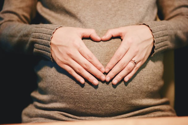 En person i brun tröja som sitter med händerna formade som ett hjärta över sin gravidmage