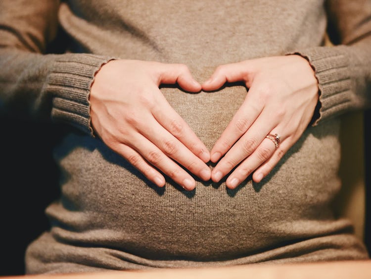En person i brun tröja som sitter med händerna formade som ett hjärta över sin gravidmage