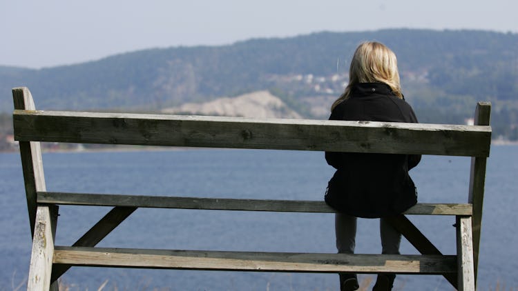 En person sitter ensam längst ut på en bänk som är vänd mot en sjö.