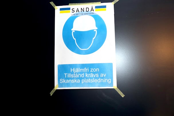 En utskriven skylt med en stiliserad bild på en person i bygghjälm, där det även står: SANDÅ. "Hjälmfri zon. Tillstånd krävs av Skanska platsledning".