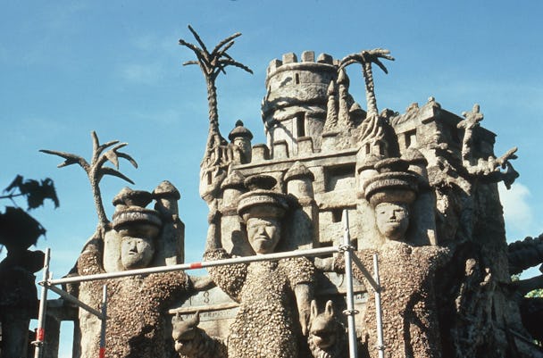 Ett sandfärgat palats med krenelerade torn. Framför byggnaden står tre människoskulpturer i samma färg.