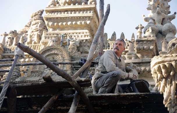 En man med säkerhetslina sitter på huk med händerna ovanför en hink, framför ett sandfärgat palats.