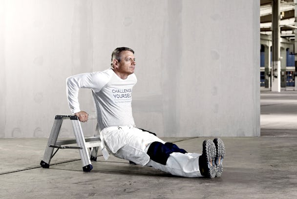 En man sitter på golvet framför en stegpall, och lyfter upp sin kropp med hjälp av händerna mot pallens överdel.
