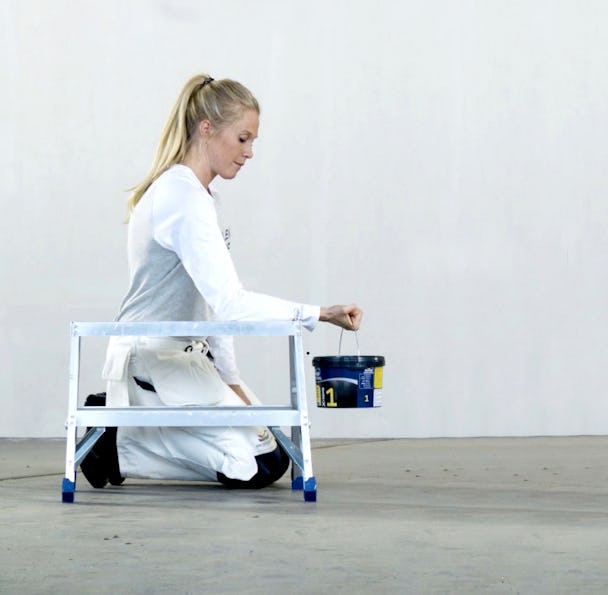 En kvinna sitter på knä på golvet med en färghink i händerna.