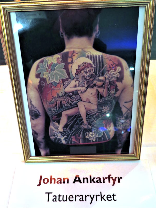 En ryggtavla med en stor tatuering föreställande en naken man som spelar fiol i ett vattendrag.