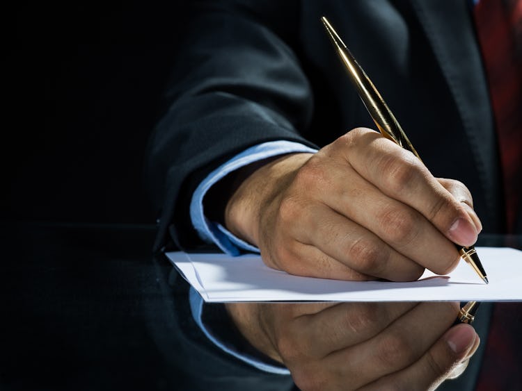 Handen på en person i kostym som skriver på ett papper med en fin penna