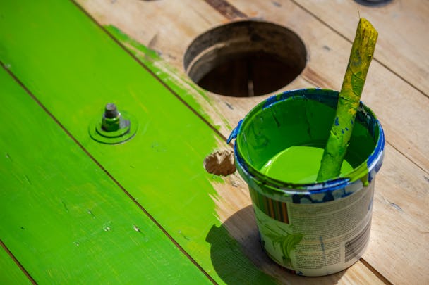 En hink med grön färg samt pensel på ett halvt målat golv