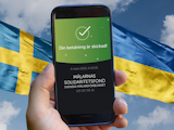 En mobiltelefon som visar en godkäns swish-betalning till Målarnas solidaritetsfond, framför en bild på Sveriges och Ukrainas flaggor och en blå himmel.