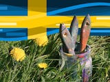 Flera penslar i en burk, burken står bland högt gräs, ovanför svajar en svensk flagga.