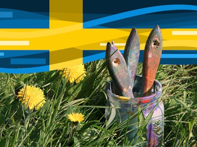 Flera penslar i en burk, burken står bland högt gräs, ovanför dom svajar en svensk flagga.