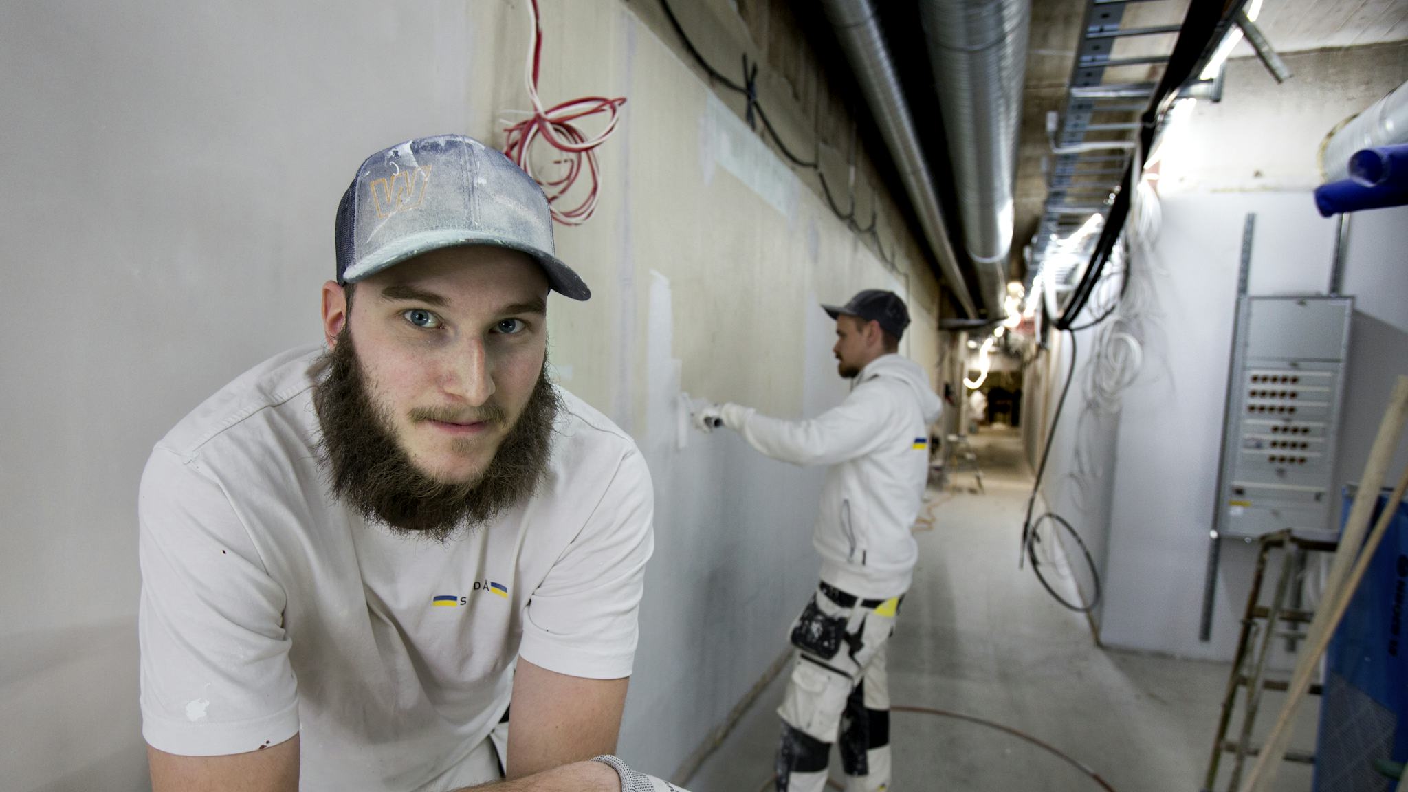 Ludwigh Eriksson Melin tittar in i kameran, Conny Forsberg står bakom och målar en vägg.