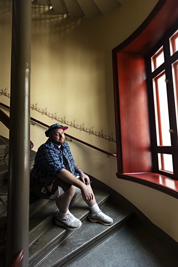 Målaren Miku Godfrey sitter på en trappa nära ett fönster och tittar mot ljuset som strömmar genom det öppna fönstret.
