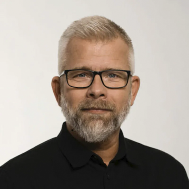 joakim nexbo med grått hår och skägg, iförd glasögon och en svart skjorta, blickar mot kameran framför en enhetlig vit bakgrund.