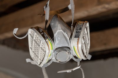 En andningsmask med dubbelt filter upphängd i sina remmar i en verkstad, med en träbjälke i bakgrunden.