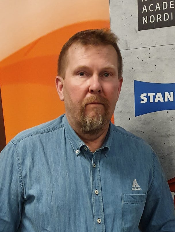 Anders Helgesson med skägg som står framför en vägg med reklambanderoller, klädd i blå jeansskjorta.